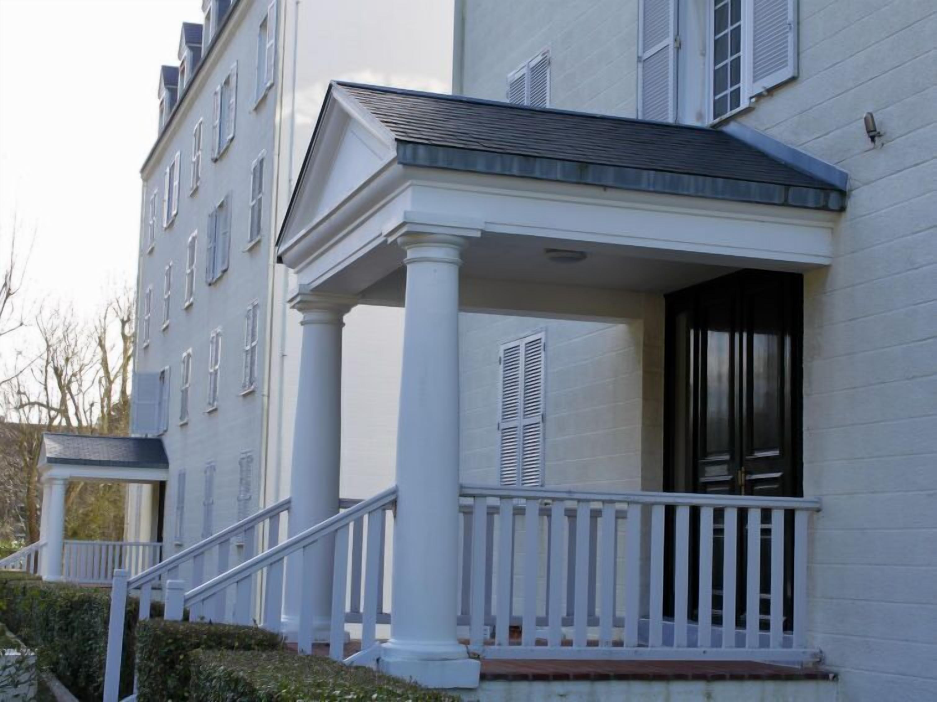 Un edificio blanco con un porche sostenido por columnas, una escalera y una puerta con contraventanas.