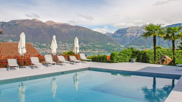 Ein Pool mit weißen Liegestühlen und Sonnenschirmen, umgeben von Grünpflanzen und Bergen im Hintergrund.