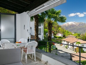 A balcony with white chairs and a table, overlooking a mountainous landscape with houses and palm trees. - Residenza Miralago Apt. A8 by Interhome (Piazzogna)