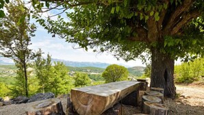 Uma área de estar ao ar livre rústica, com uma longa mesa de madeira e bancos feitos de tocos, emoldurada por paisagem natural, árvores e vista para colinas distantes.