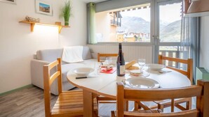 Un coin repas avec une table ronde, des chaises en bois et un canapé blanc. Il y a une fenêtre donnant sur les montagnes et un balcon.