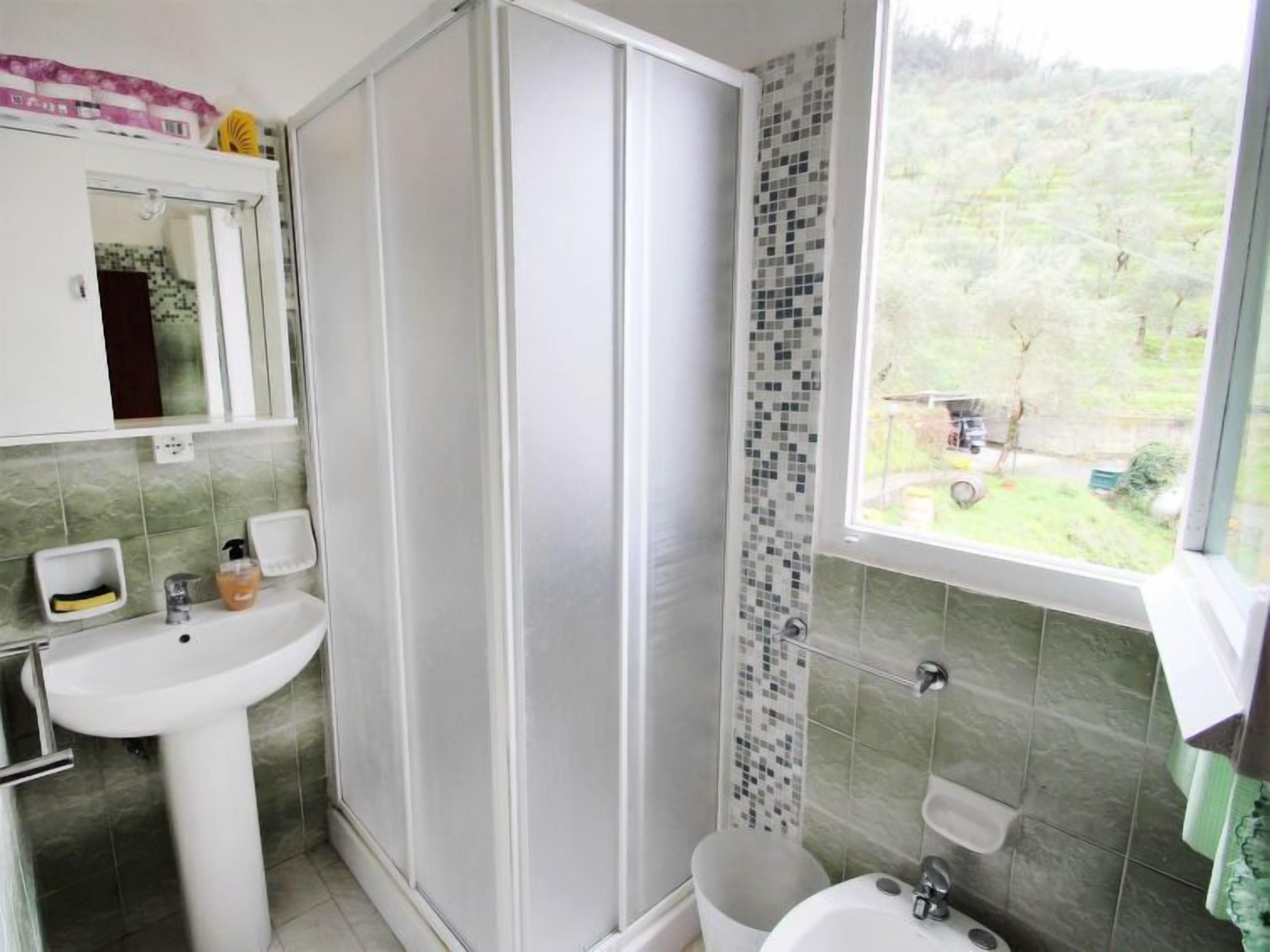 Ein Badezimmer mit Dusche, Waschbecken und Toilette. Ein Fenster bietet Blick auf Grünfläche außerhalb.