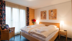 Ein Hotelzimmer mit einem großen Bett, zwei Nachttischlampen, einem Sessel mit floralem Kissen und einer Vase mit roten Blumen.