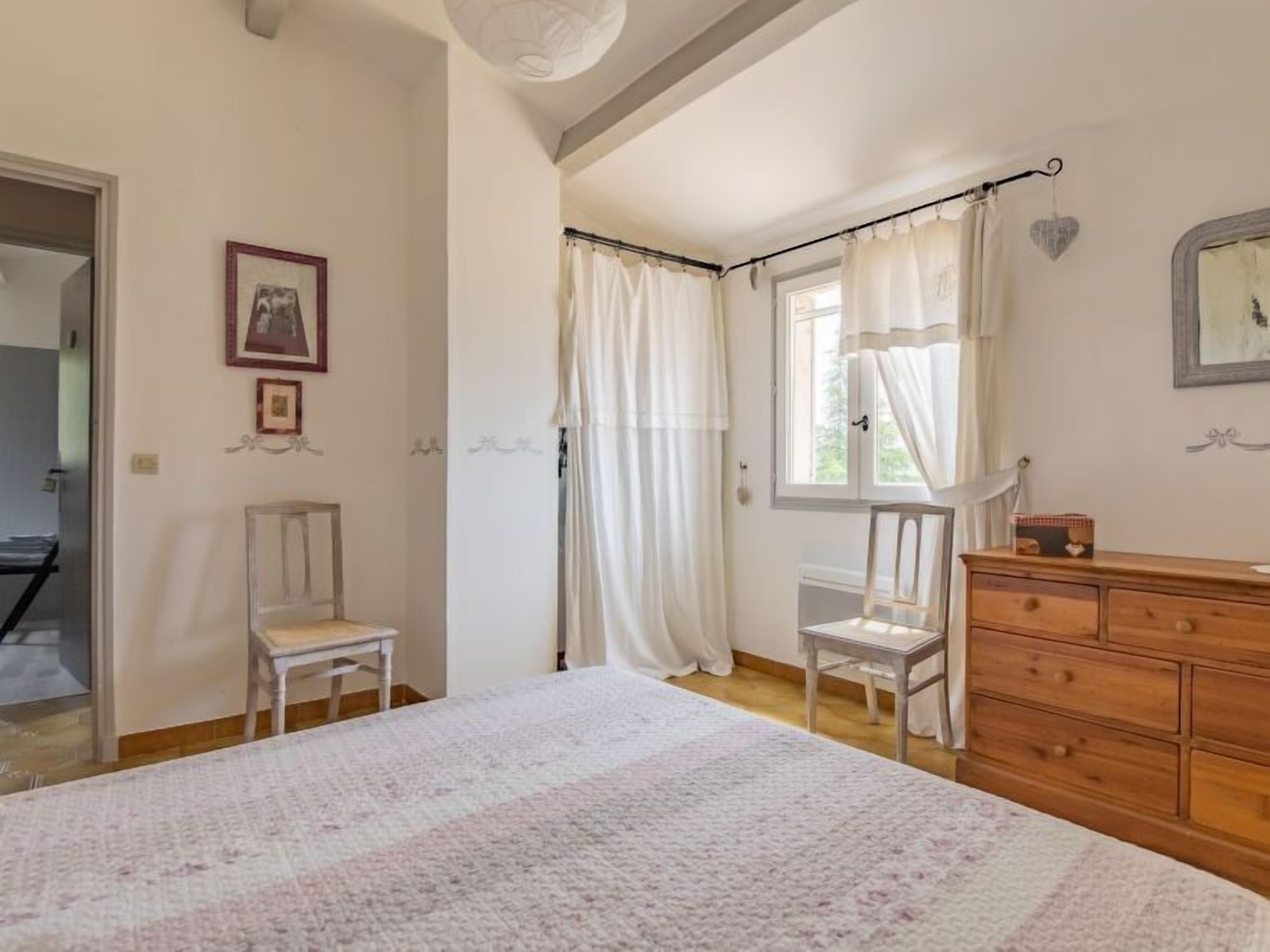 A bedroom with a bed, two chairs, a dresser, and a window with curtains.