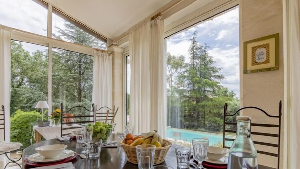 Ein Esstisch ist gedeckt mit Tellern, Schüsseln, Gläsern und einer Flasche, mit Blick auf einen Pool und Bäume im Hintergrund.