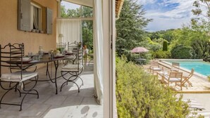 Eine überdachte Terrasse mit Esstisch und Stühlen, ein Poolbereich mit Liegestühlen und Blick auf die umliegende Grünfläche.