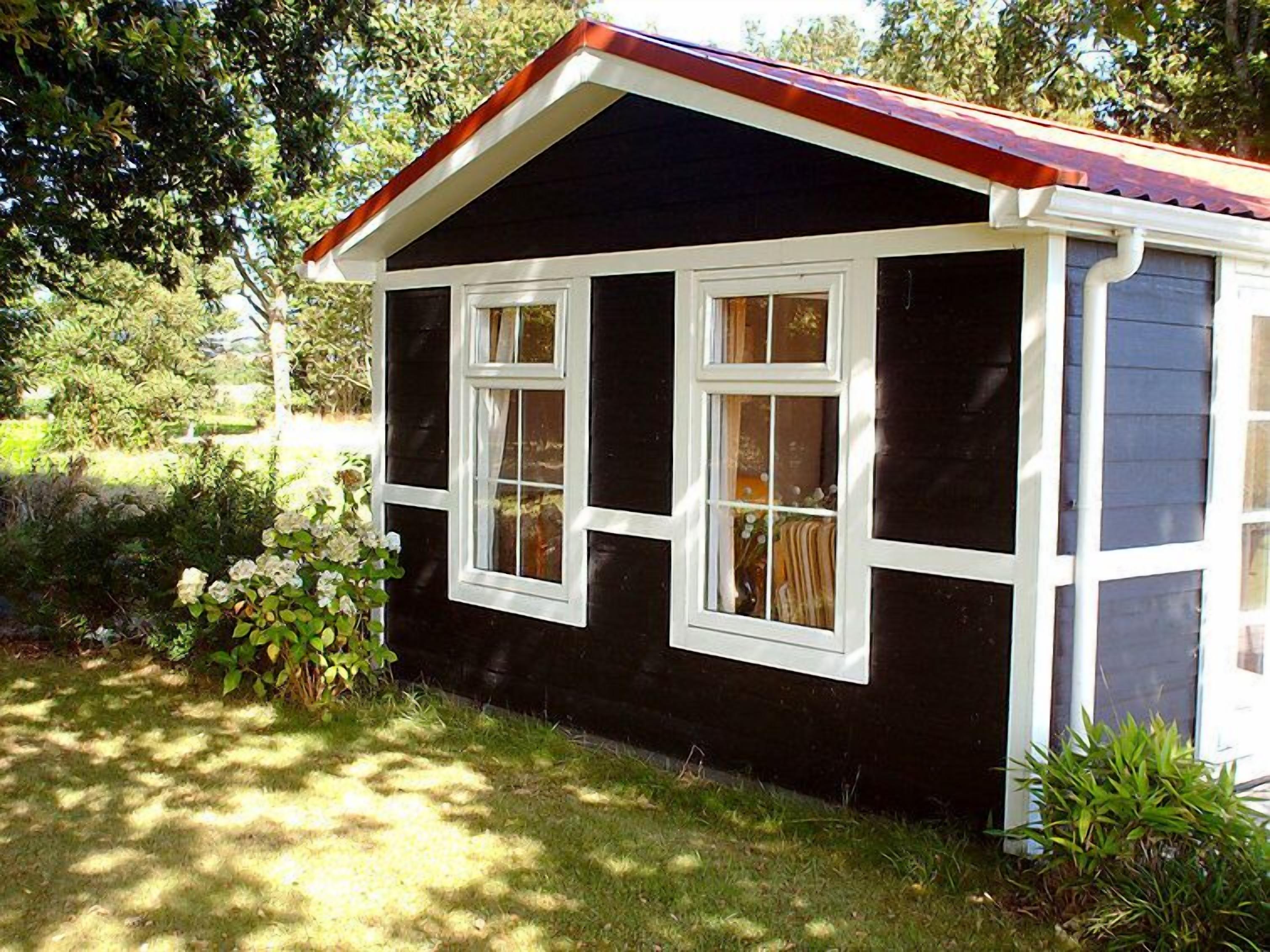 Una casa pequeña de color oscuro con techo rojo y detalles blancos. Tiene varias ventanas y está rodeada de follaje.