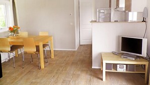 Ein modernes, gut beleuchtetes Zimmer mit einem Holzespeisebereich, Sesseln, einem Fernseher auf einem Schrank und einer Küchenzeile mit Geräten.