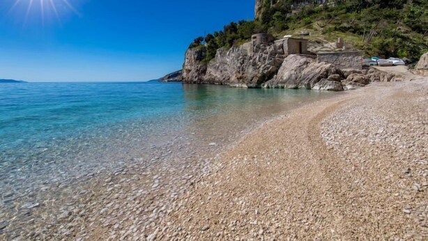 Une plage de galets bordée d’une eau claire, une falaise rocheuse et un bâtiment perché au sommet.