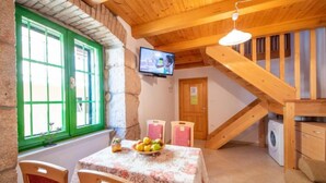 Une pièce chaleureuse avec un plafond en bois, un mur en pierre, une fenêtre ornée d’un cadre vert, une table avec des fruits et un lave-linge.