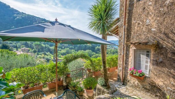 A patio with a table set for two, surrounded by potted plants and a large umbrella. - Il Metatino by Interhome (Camaiore)