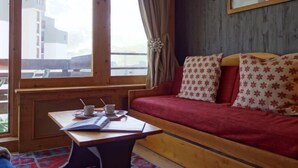 Ein gemütliches Zimmer mit einer Holzcouch, einem kleinen Tisch mit Tassen und einem Buch sowie einem Fenster mit Blick auf Gebäude.