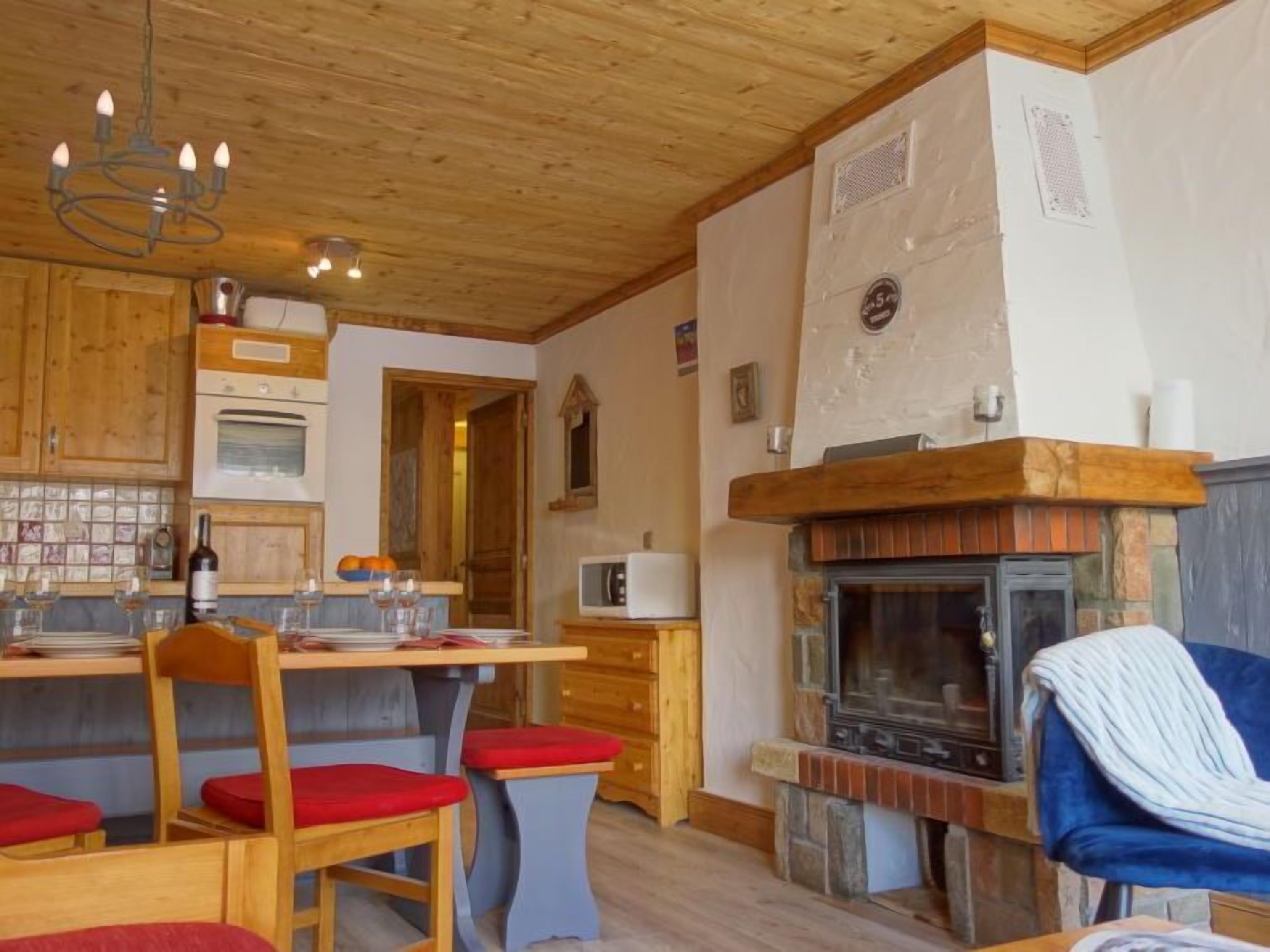 A cozy kitchen with wooden cabinets, a dining table with red cushioned chairs, a built-in stove, and a blue chair with a white throw.