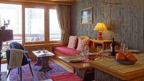 Ein gemütliches Zimmer mit Holzbalkendecke, einem Sofa mit gemusterten Kissen, einem Esstisch gedeckt mit Tellern und Gläsern sowie einem Fenster mit Vorhängen.