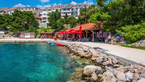 A coastal area with clear turquoise water, a rocky shoreline, and a small pier. There are buildings in the background, including a multi-story structure with balconies. - Apartment Delfin by Interhome (Karlobag)
