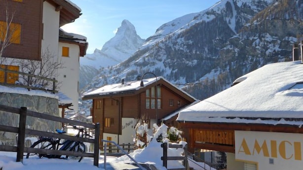 Un pueblo nevado en la montaña con edificios de madera, un pico montañoso destacado al fondo y un cartel que dice "AMICI".