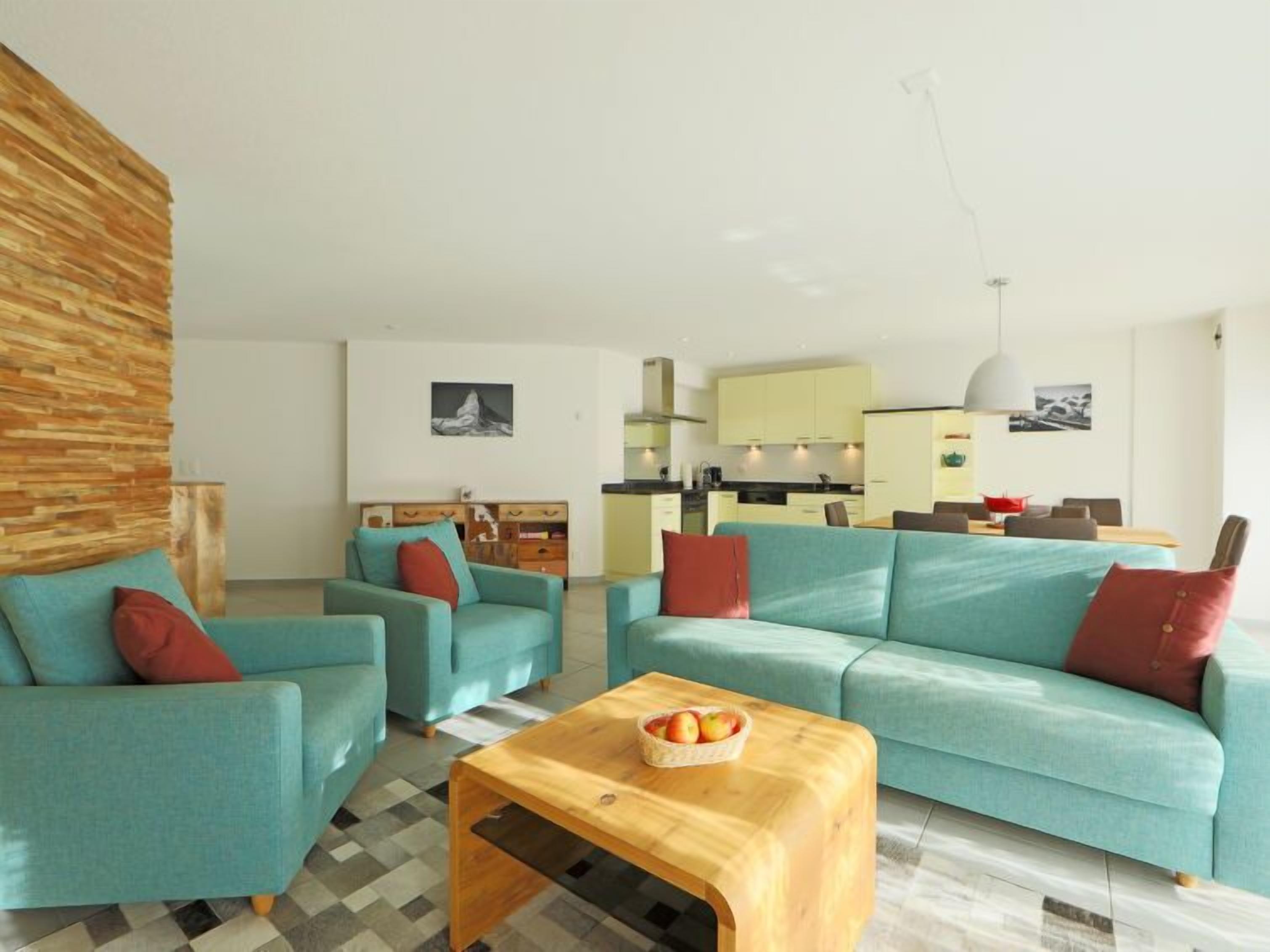 A modern living room with teal sofas, a wooden coffee table, and a kitchen in the background.