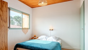 A bedroom with a wooden ceiling, a bed with a teal blanket, a nightstand with a lamp, and a window with curtains. - Vacation home Sicard by Interhome (Mimizan)
