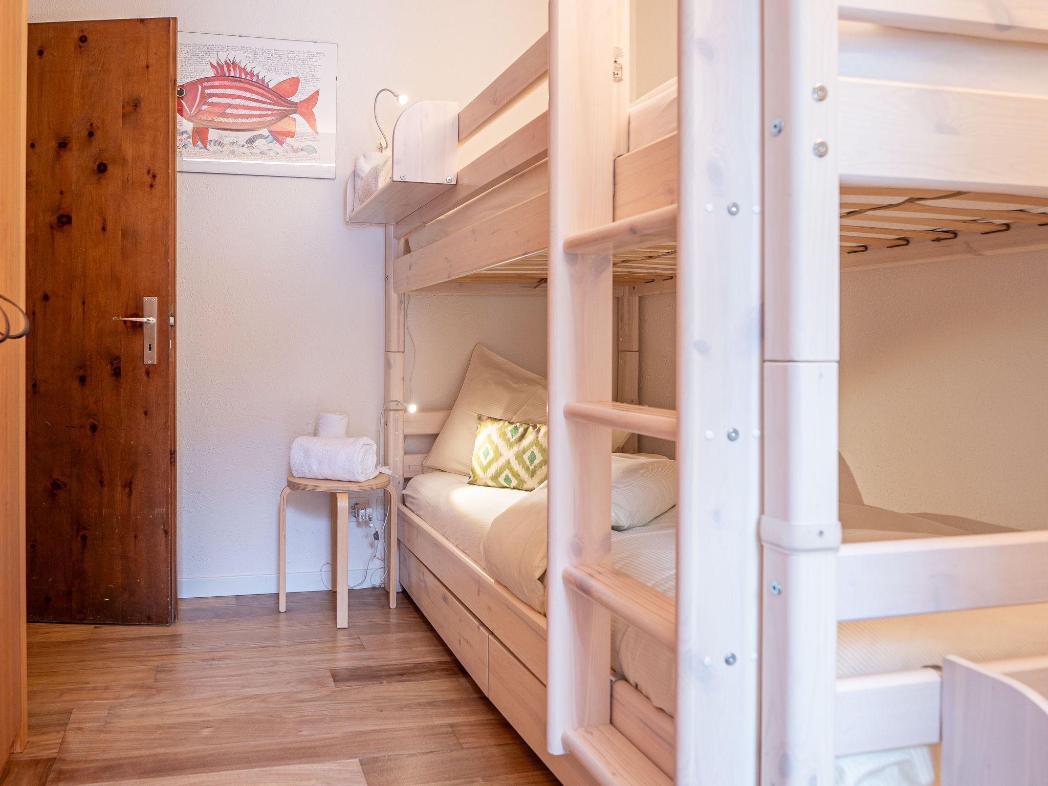 Une chambre avec des lits superposés, une porte en bois, une petite table avec une serviette et une illustration encadrée représentant un poisson accrochée au mur.