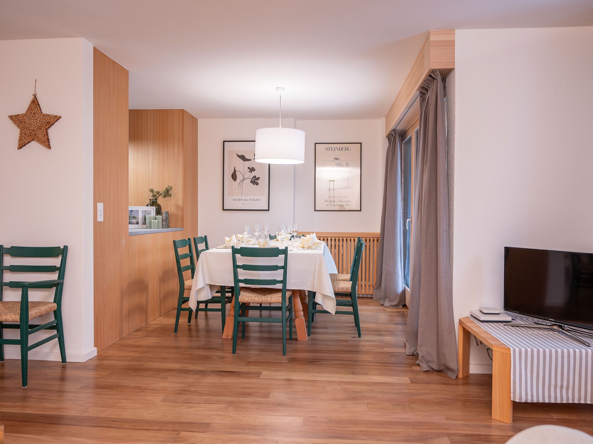 Un coin repas avec une table en bois dressée pour quatre personnes, des chaises vertes, un luminaire suspendu et un tableau représentant une fleur.