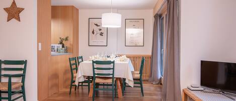 Un coin repas avec une table en bois dressée pour quatre personnes, des chaises vertes, un luminaire suspendu et un tableau représentant une fleur.