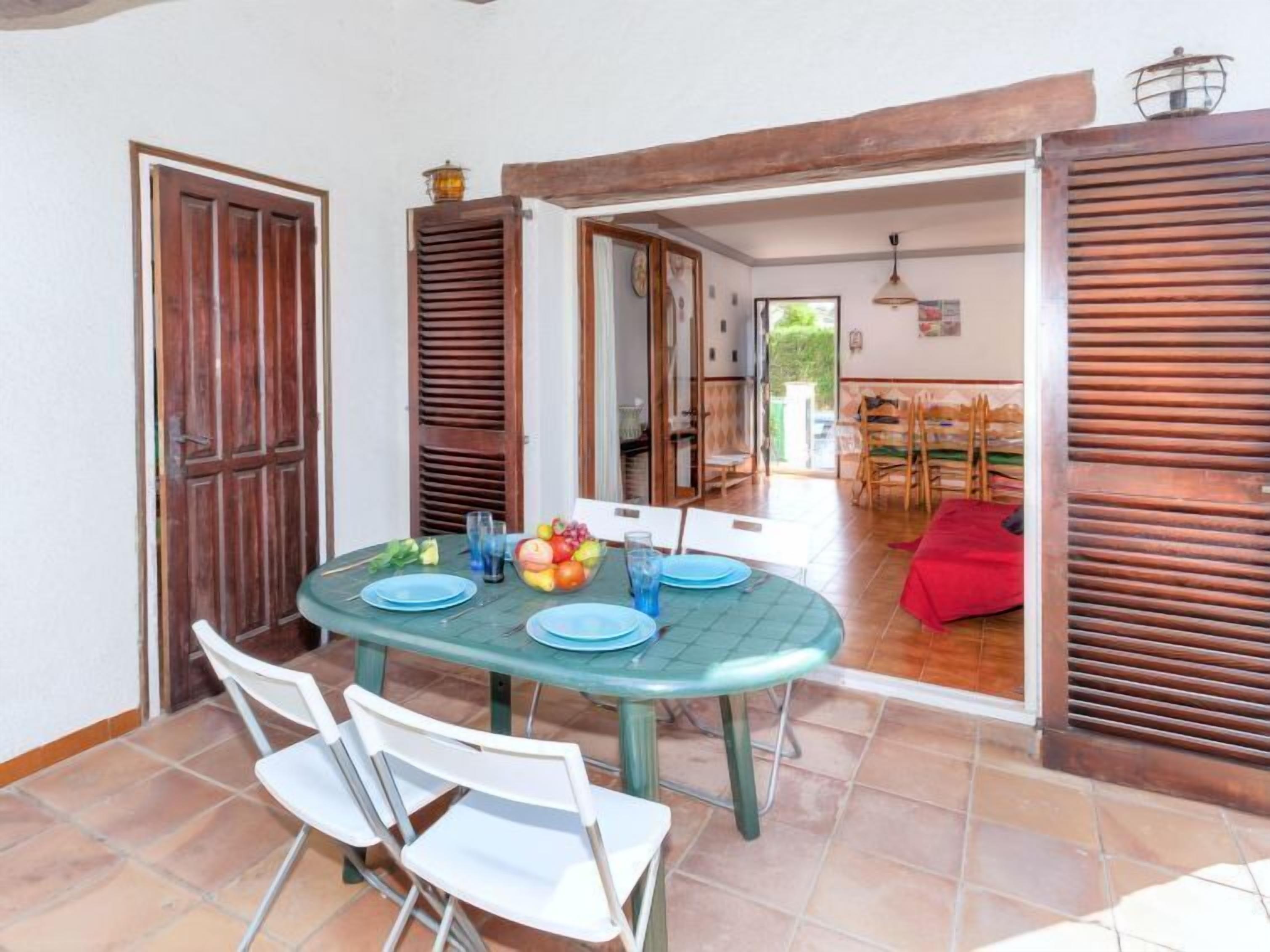 A dining area with a round table, white chairs, and a fruit bowl. There are wooden shutters on the walls and a red cushion on the floor.