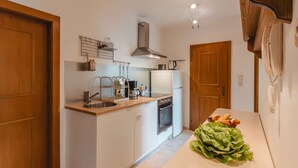 Cucina compatta con lavandino, fornelli e frigorifero. Una porta in legno a sinistra e un piano di lavoro bianco con una ciotola di verdure a destra.