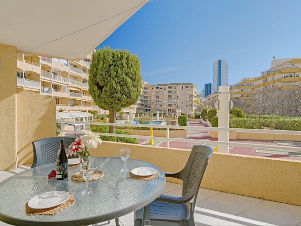 A balcony with a round glass table set for dining, a potted tree, and a view of apartment buildings and a pool.