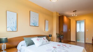 Quarto com cama de madeira, colcha florida, duas luminárias fixadas na parede e porta que dá para outro cômodo.