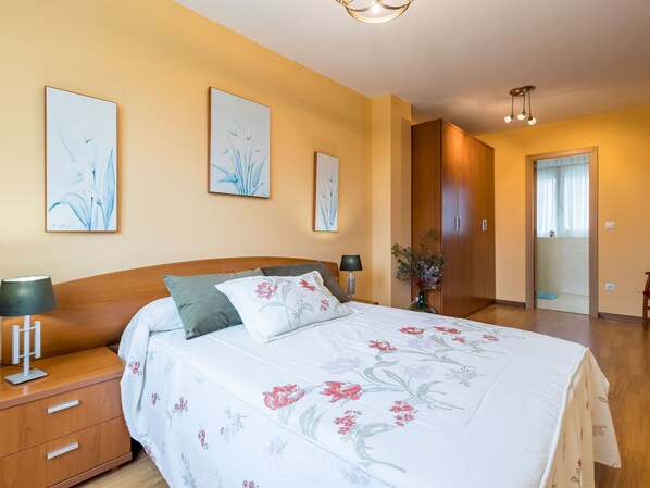 Quarto com cama de madeira, colcha florida, duas luminárias fixadas na parede e porta que dá para outro cômodo.