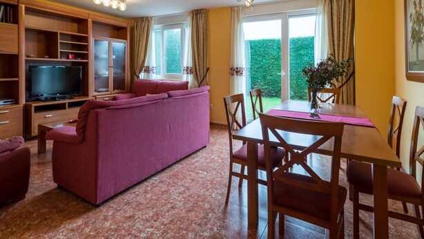 Sala de estar com sofá, televisão, prateleiras de madeira e área de jantar com cadeiras e mesa.