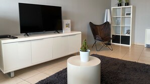 Ein modernes Wohnzimmer mit einem Flachbildfernseher auf einem weißen Schrank, einem dunkelbraunen Ledersessel, einem kleinen weißen Tisch mit Pflanze und einer weißen Regaleinheit mit Pflanzen und Dekorationsgegenständen.