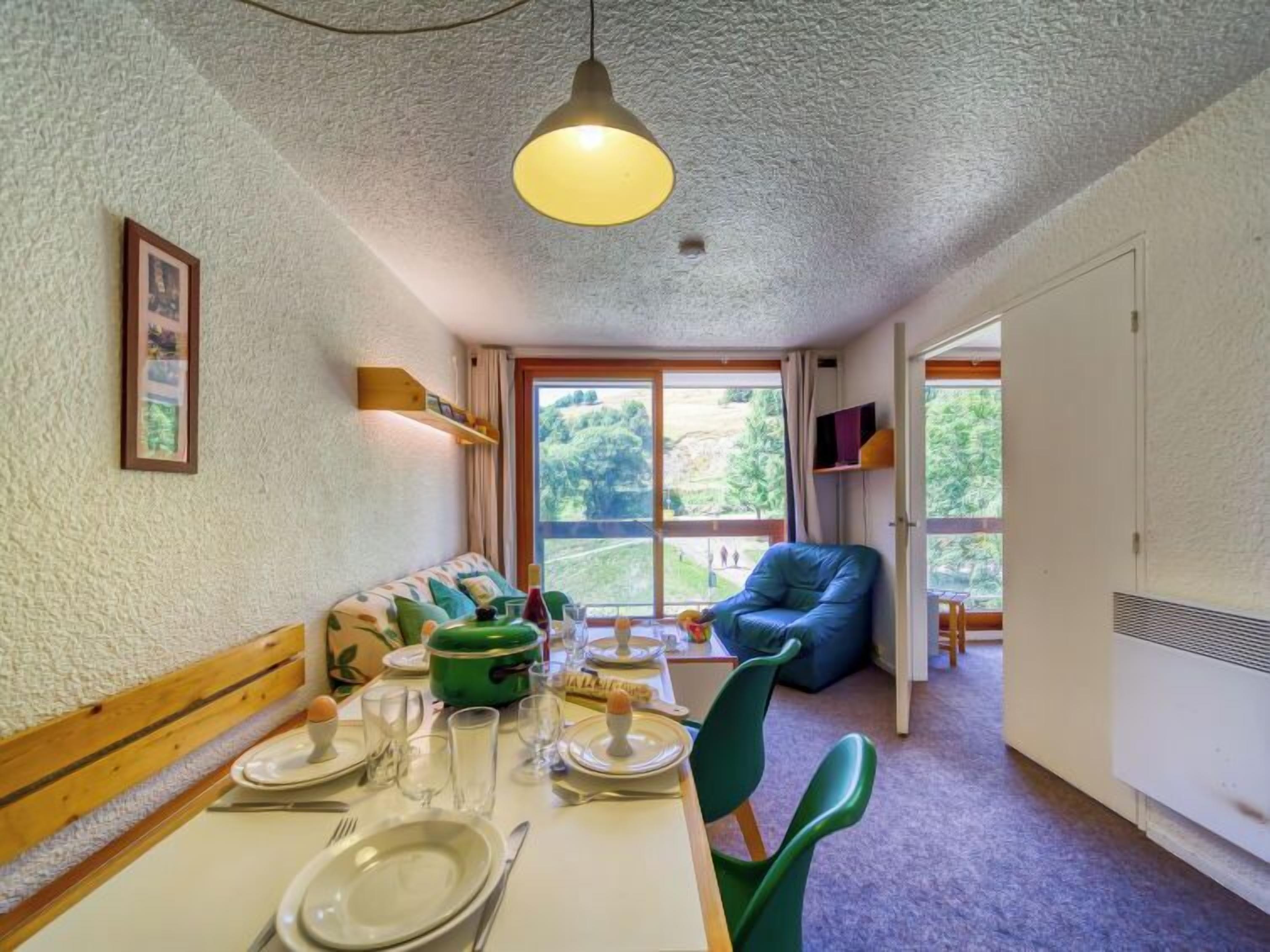 Un comedor con una mesa preparada para cuatro personas, un sofá verde, una silla azul y un ventanal con vistas a zonas verdes.