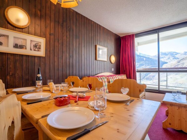 A cozy room with a wooden table set for dining, a bed with a colorful blanket, and a large window offering a mountain view.