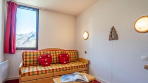 A cozy room with a wooden sofa, red cushions, a small wooden table with a magazine, a window with red curtains, and a wall-mounted light fixture.