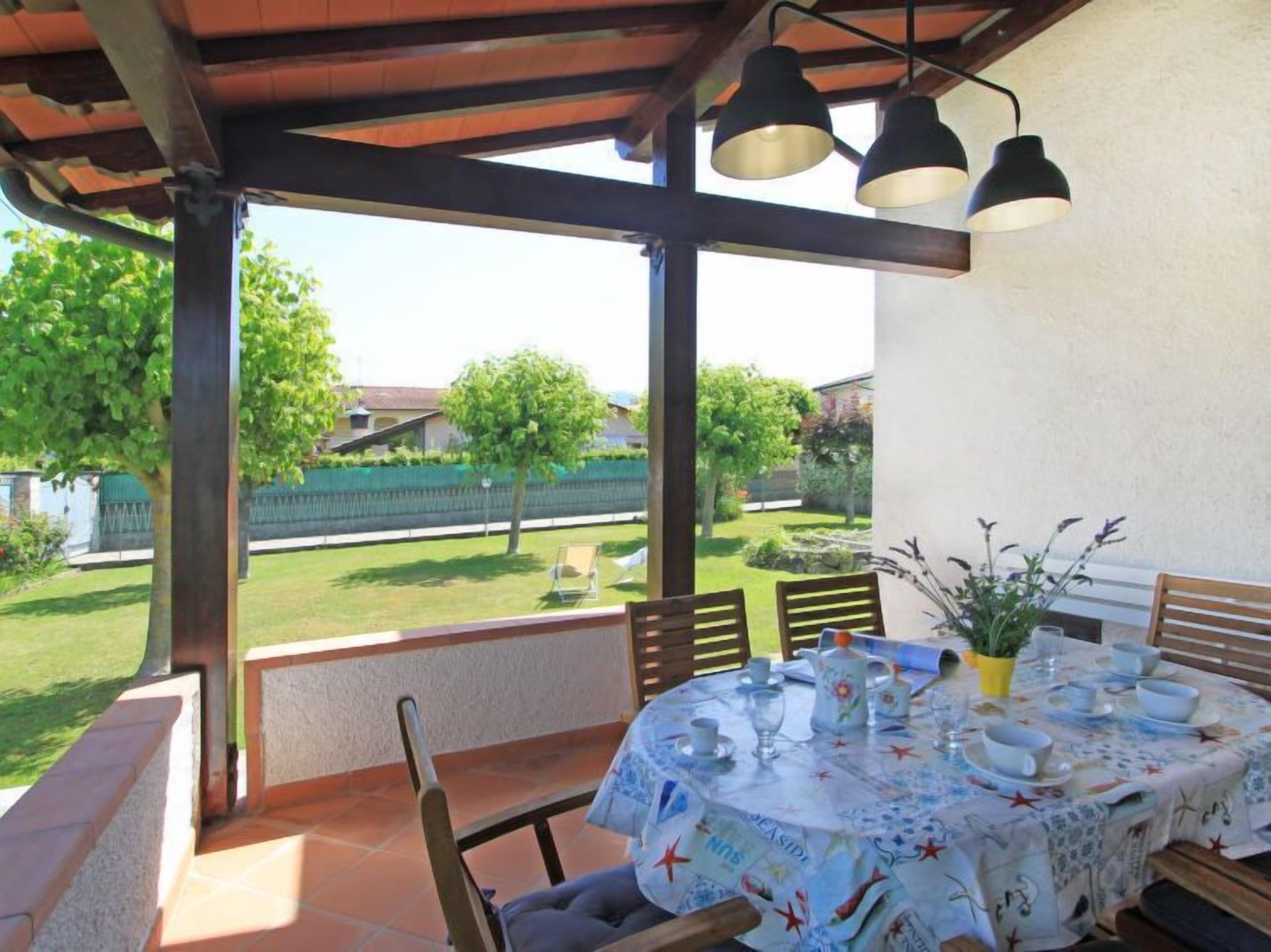 An outdoor patio with a table set for dining, surrounded by chairs, under a wooden pergola with hanging lights.