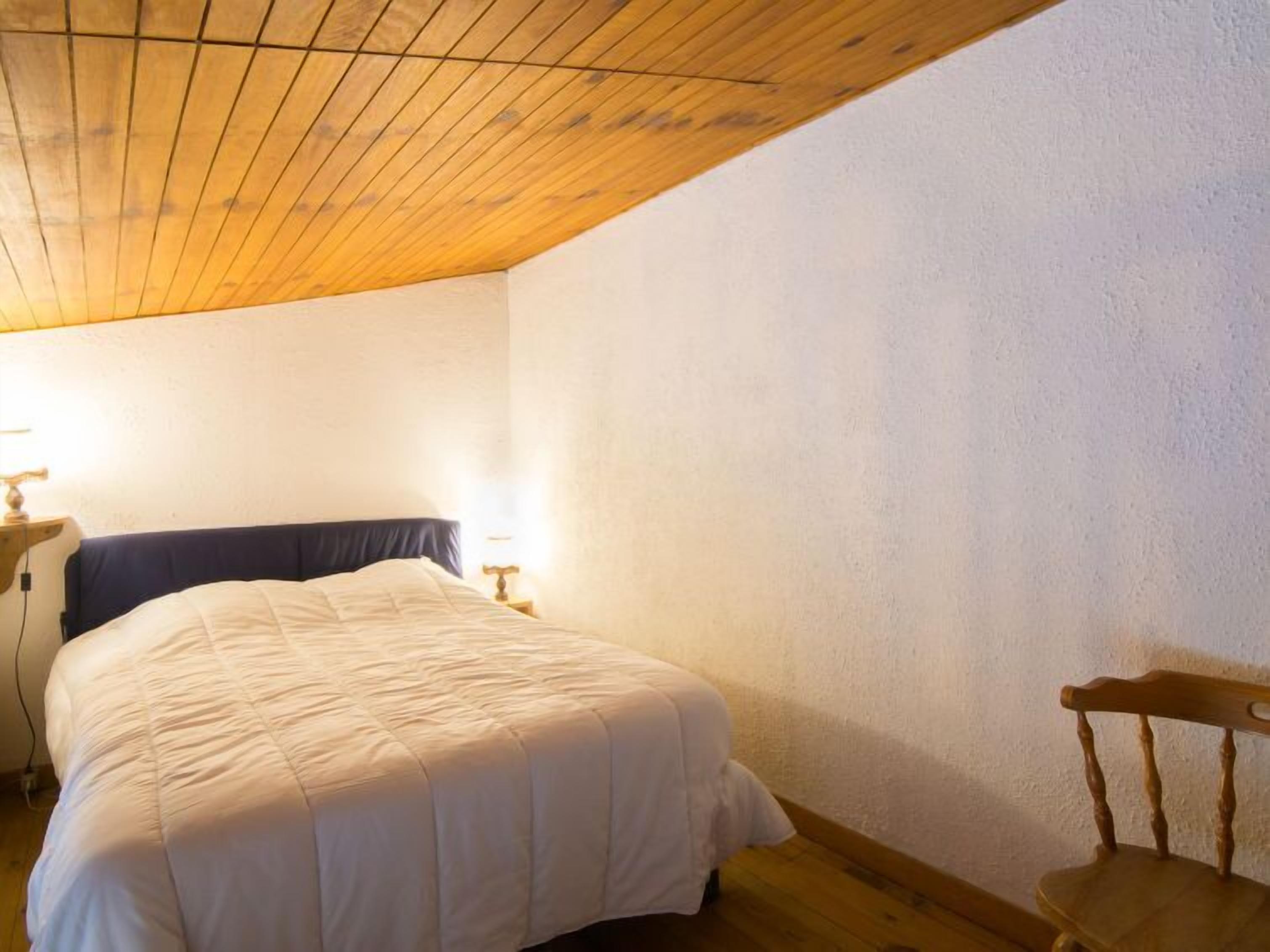 Ein Schlafzimmer mit Holzbalkendecke, einem Bett mit weißer Daunendecke, zwei Nachttischlampen und einem Holzstuhl.