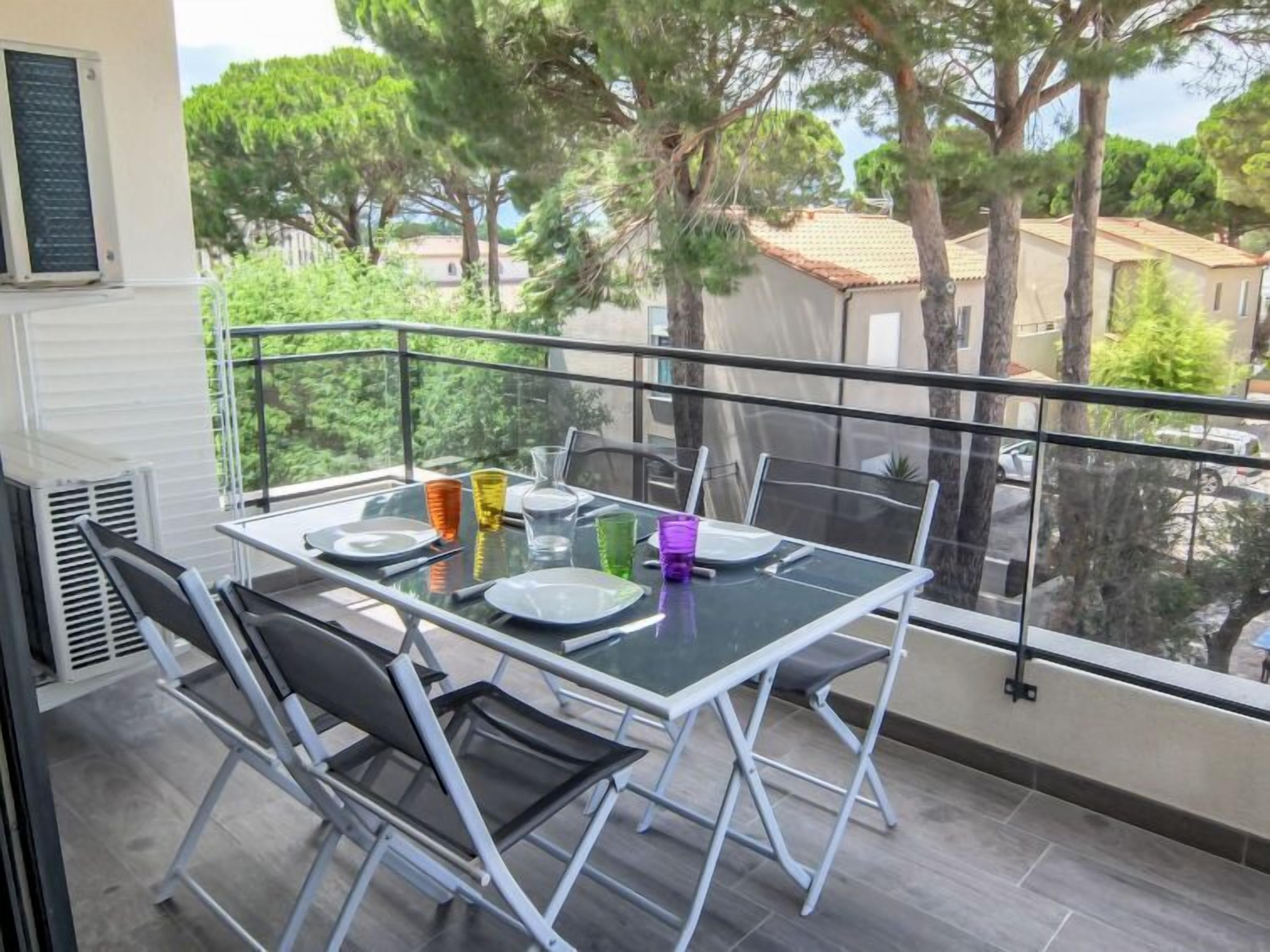 Un balcón con mesa de vidrio dispuesta para comer, rodeado de sillas metálicas, con vista a una zona residencial con árboles y casas.