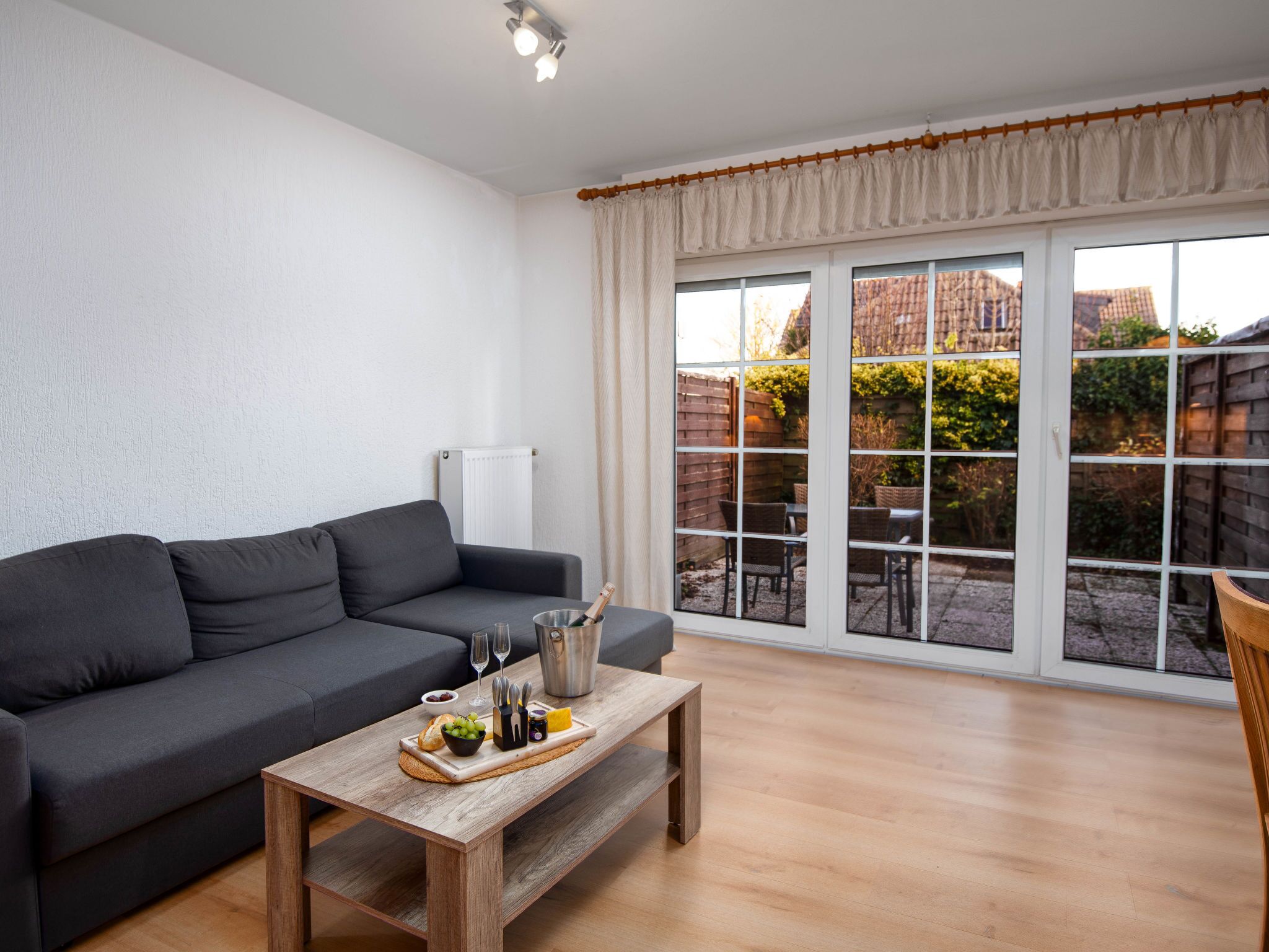A modern living room with a grey sofa, wooden coffee table with food and drinks, and large windows overlooking a garden.
