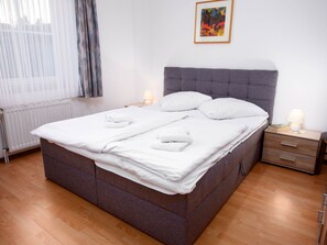 Un lit bien fait, avec du linge de lit blanc, une tête de lit rembourrée grise et deux tables de chevet avec des lampes.