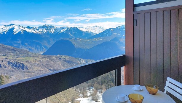 Varanda com mesa e cadeiras, com vista para uma paisagem montanhosa com picos nevados.
