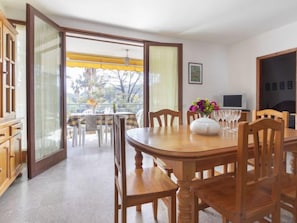 Een eetruimte met een houten tafel en stoelen, een witte vaas met bloemen, en uitzicht op het buiten via een glazen deur.