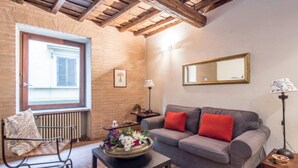 Ein gemütliches Wohnzimmer mit grauem Sofa, roten Kissen, einem schwarzen Metalltisch mit Blumenarrangement, einer Holzbank mit gemusterten Kissen, einer Heizung, einem Fenster mit Rollläden und einem Spiegel an der Wand.