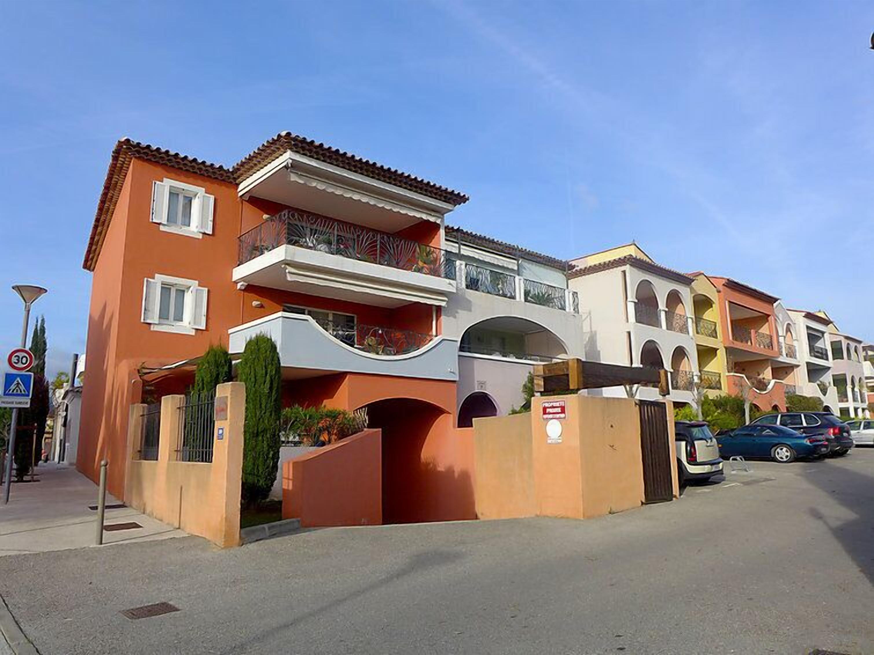 Un edificio residencial de varios pisos con balcones, un acceso con portón y un cielo azul despejado.