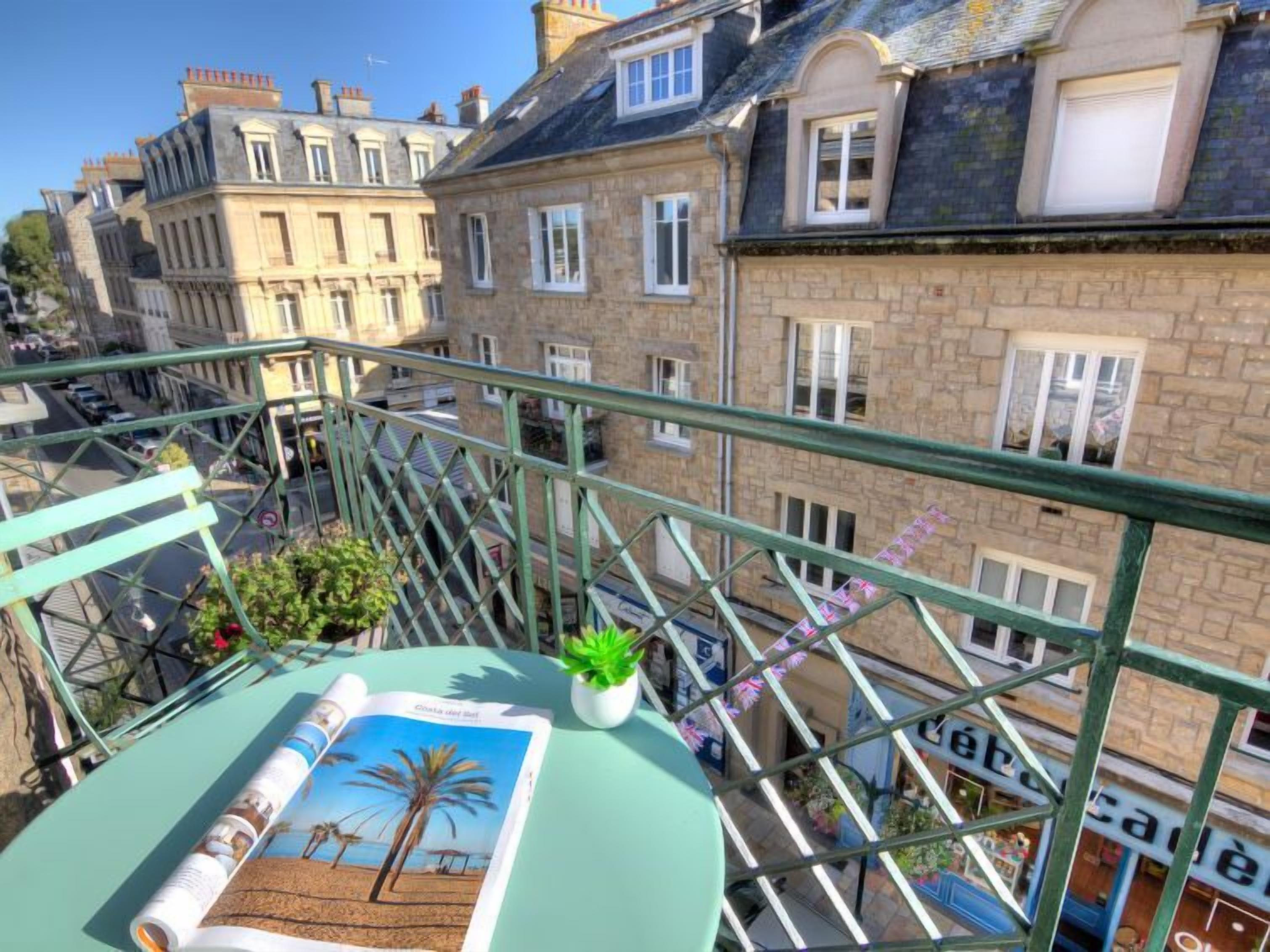 A balcony with a view of old stone buildings, a small table with a magazine and a plant, and a railing.