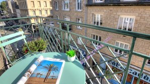 Un balcón con vistas a edificios de piedra antiguos, una mesita con una revista y una planta, y una barandilla.