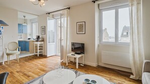 Una sala de estar moderna con televisor, una mesa de comedor con copas de vino, una zona de cocina con armarios azules y ventanas amplias con cortinas.