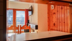 Una habitación con una mesa de comedor de madera, sillas, un televisor montado en la pared y una ventana con cortinas rojas.