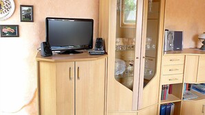 Una stanza con un armadio in legno che contiene una TV a schermo piatto, un altoparlante e un telecomando. Alle pareti ci sono quadri incorniciati e una libreria piena di libri.
