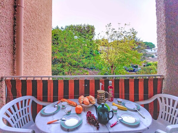 Un balcon avec une table dressée pour deux personnes, comprenant des assiettes, des tasses, un pichet, une théière et une bouteille.